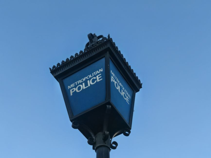 Close-up view of a vintage-style street lamp with a square blue sign displaying the words 'Metropolitan Police' on each side, mounted on a black metal pole with decorative scrollwork near the top, set