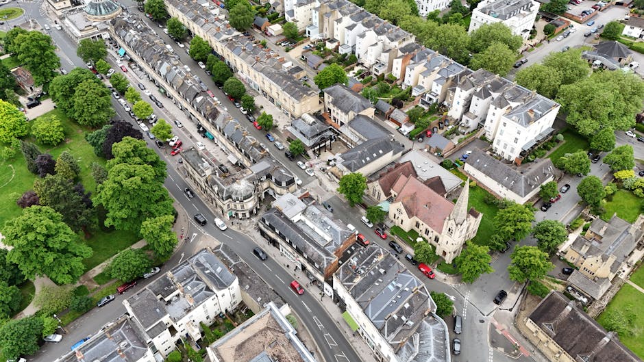 An aerial view of Ealing Broadway showing a narrow street with parked cars along both sides, surrounded by residential buildings and green trees. Visible are various vehicles including vans and cars, some positioned near the curbs, with a church with a tall spire also situated along the street. The scene captures the typical urban environment during daylight, with natural light illuminating the scene. This image illustrates the urban landscape common in areas requiring house removals or furniture transport, as carried out by services such as those provided by Ealing Man and Van, particularly in streets with tight access for loading and unloading during home relocations.
