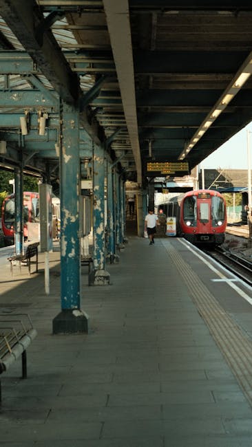 Inside a train station with a platform on the left side and railway tracks extending into the distance. The platform has concrete paving and a metal railing acting as a safety barrier. A moving train