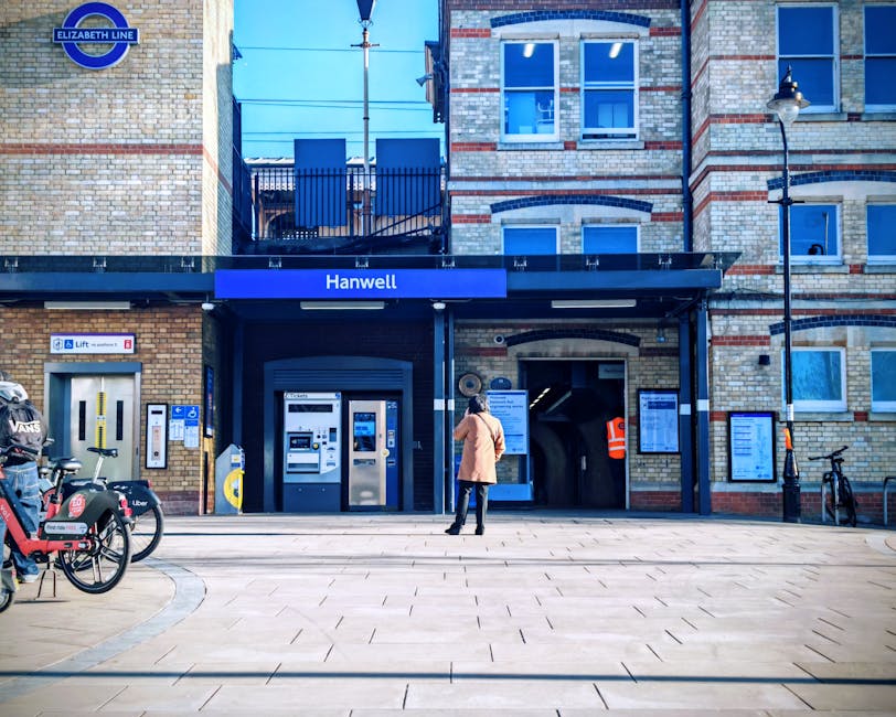 A woman dressed in a beige coat and black trousers stands outside the entrance of Hanwell station, part of the Elizabeth Line, in the W5 postcode area, with the station sign visible above her. The station building features a brick facade with a blue canopy over the entrance. To the left, there are several bicycles parked, including one with a container marked for a delivery service, and a row of ticket vending machines and automated barriers are positioned along the pavement. To the right, a bicycle stands next to a black lamppost, and a person in an orange high-visibility vest is visible near the station entrance, possibly involved in the loading or unloading process. The scene is illuminated by natural daylight, casting soft shadows on the tiled pavement, suggesting a clear day. This setting may relate to home relocation or moving services, highlighting the transportation and logistical aspects of a house or flat move, with a focus on the station's entrance and surrounding environment, as seen in the context of the 'W5 postcode man and van tips for flats above shops, EALING' page on ealingmanandvan.net.